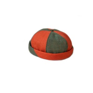 Könnte beinhalten: Ein zweifarbiges Hut in einem klassischen Stil. Der Hut ist in orangefarbene und olivgrüne Abschnitte unterteilt. Der Rand ist orange und die Krone ist eine Kombination aus den beiden Farben. Der Hut ist aus einem strukturierten Stoff gefertigt.