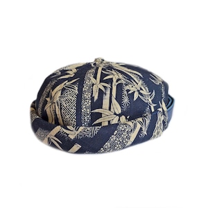 May include: A navy blue skull cap featuring a beige bamboo and geometric design. The cap has a short brim and a rounded crown. The fabric has a textured appearance, and it is set against a white background.