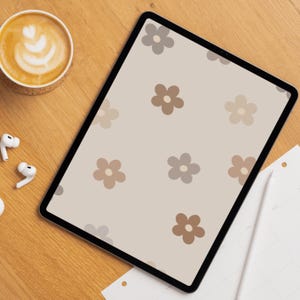 Op de afbeelding: Een tablet met een digitale achtergrond met een herhalend patroon van eenvoudige vijfbladige bloemen in tinten beige, lichtbruin en bruin op een crèmekleurige achtergrond. De tablet ligt op een houten oppervlak met koffie en oordopjes.