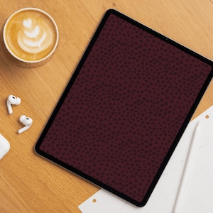 Op de afbeelding: Een digitale tablet met een bordeauxrode stippenpatroon op het scherm ligt op een houten oppervlak. Een kopje koffie met latte art, witte oortelefoons en een stylus zijn ook zichtbaar. De scène suggereert een werkplek of een moment van ontspanning.
