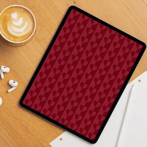 Op de afbeelding: Een digitale tablet met een dieprode achtergrond met een herhalend verticaal patroon van donkerrode harten gerangschikt in een rankachtige volgorde. Het scherm is omlijst door een dunne zwarte rand en ligt op een lichtgekleurd houten oppervlak.