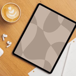 Op de afbeelding: Een digitale tablet met een geometrisch patroon in bruintinten, een kopje koffie met latte art, draadloze oortelefoons en een notitieblok op een houten oppervlak. Het scherm van de tablet toont een modern, abstract ontwerp.