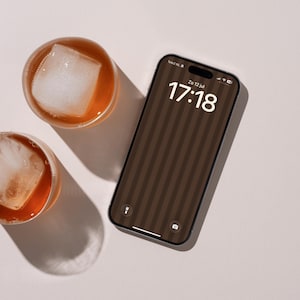 Op de afbeelding: Een smartphone met een donkerbruine verticaal gestreepte achtergrond die de tijd 17:18 en de datum Zo 12 jul weergeeft, naast twee ronde glazen gevuld met amberkleurige vloeistof en grote vierkante ijsblokjes.