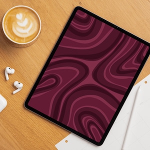 Op de afbeelding: Een digitale tablet met een abstract, golvend patroon in bordeauxrood. Een kopje koffie met latte art, draadloze oortelefoons en een notitieblok liggen op een houten oppervlak.