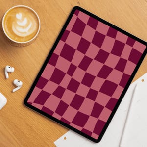 Op de afbeelding: Een digitale tablet met een bordeauxrood en roze ruitpatroon op het scherm. Een kopje koffie met latte art, witte draadloze oortelefoons en een witte stylus liggen op een houten oppervlak.