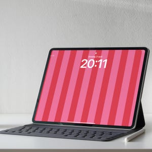 Op de afbeelding: Een zwarte tablet met een toetsenbordhoes en stylus. Het scherm toont een roze en rode streepjespatroon met de tijd 20:11. De tablet staat op een wit oppervlak.