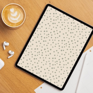 Op de afbeelding: Een digitale tablet met een crèmekleurig scherm met een patroon van kleine, lichtgroene stippen. Een kopje koffie met latte art, draadloze oordopjes en een notitieblok liggen op een houten oppervlak.