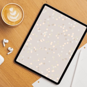 Op de afbeelding: Een digitale tablet met een zwarte rand toont een beige achtergrond met een patroon van kleine, lichtgekleurde cirkels. Een kopje koffie met latte art, draadloze oortelefoons en een notitieblok met een stylus liggen op een houten oppervlak.