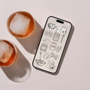 Op de afbeelding: Een smartphone met een vergrendelscherm waarop een raster van zwarte lijntekeningen van bak-iconen staat, waaronder een kom, brood, een pot, oven, meelzak, tarwe, garde, ovenwanten en een mixer, naast twee glazen met een amberkleurige drank.