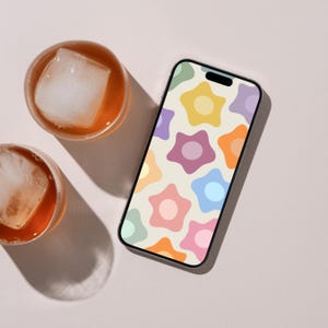 Op de afbeelding: Een smartphone met een scherm dat een herhalend patroon van veelkleurige, vijfbladige bloemen in tinten geel, oranje, roze, blauw en paars op een crèmekleurige achtergrond toont, naast twee glazen ijsthee met ijsblokjes.