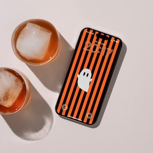 Op de afbeelding: Een smartphone met een vergrendelscherm met een verticaal oranje en zwart gestreept patroon en een witte spookafbeelding. De tijd 20:17 is bovenaan zichtbaar. Twee ronde glazen gevuld met amberkleurige vloeistof en grote vierkante ijsblokjes.