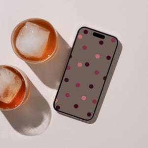 Op de afbeelding: Een smartphone met een taupekleurig scherm met een verspreid patroon van donkerpaarse en lichtroze stippen, naast twee ronde glazen gevuld met amberkleurige vloeistof en grote vierkante ijsblokjes.