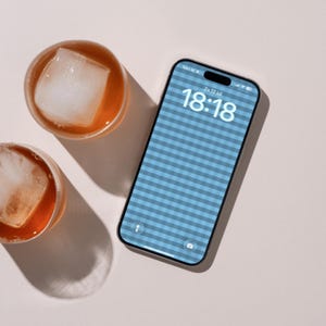 Op de afbeelding: Een smartphone met een blauw-wit vichy-ruitpatroon op het vergrendelscherm, waarop de tijd 18:18 en de datum Zo 12 jul te zien zijn. Naast de telefoon staan twee ronde glazen gevuld met amberkleurige vloeistof en grote vierkante ijsblokjes.