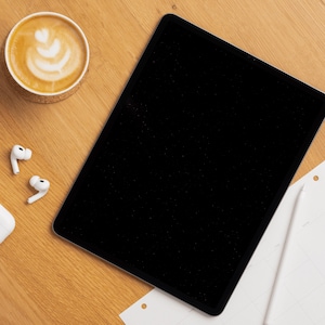 Op de afbeelding: Een zwarte tablet met een leeg scherm ligt op een houten oppervlak, naast een kopje koffie met latte art, een paar witte draadloze oortelefoons en een witte styluspen op een stuk papier.