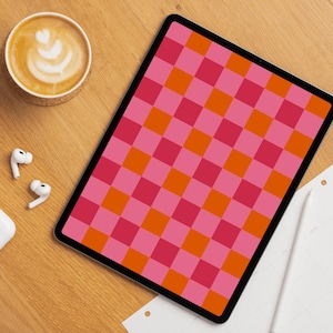 Può includere: Un tablet digitale mostra un motivo a scacchiera rosa e arancione. Una tazza di caffè con latte art, auricolari wireless e una penna stilo sono su una superficie di legno.
