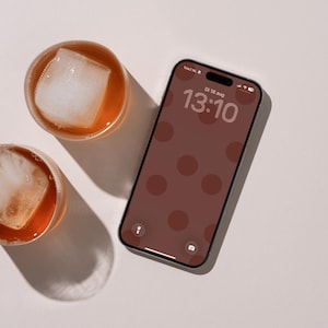 Op de afbeelding: Een smartphone met een donkerbruin scherm met een patroon van lichtbruine cirkels, waarop de tijd 13:10 en de datum Di 18 aug wordt weergegeven. Twee ronde glazen gevuld met amberkleurige vloeistof en grote vierkante ijsblokjes.