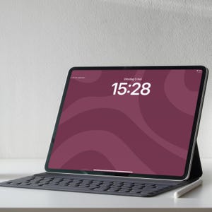 Op de afbeelding: Een zwarte tablet met een donker bordeauxrood scherm waarop de tijd, 15:28, en de datum, 5 mei, worden weergegeven. De tablet wordt ondersteund door een zwarte toetsenbordhoes en een witte stylus. De achtergrond is effen wit.