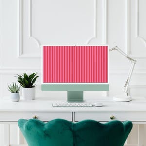 Pode incluir: Um monitor de computador de secretária com um ecrã de riscas verticais cor-de-rosa e vermelhas, com uma base verde-sálvia, colocado numa secretária branca com um teclado branco, um rato de computador branco e duas plantas verdes em vasos.