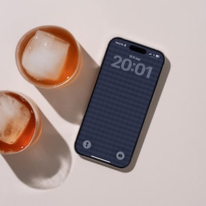 Op de afbeelding: Een smartphone met een donker scherm waarop de tijd 20:01 en de datum Di 8 sep wordt weergegeven, naast twee ronde glazen gevuld met een amberkleurige vloeistof en grote vierkante ijsblokjes, op een neutrale lichte achtergrond.
