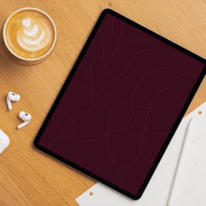 Op de afbeelding: Een digitale tablet met een bordeauxrood scherm met abstracte lijnen, een kopje koffie met latte art, draadloze oortelefoons en een notitieblok met een stylus op een houten oppervlak.