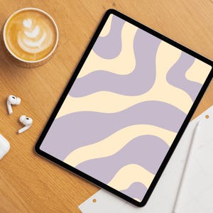 Op de afbeelding: Een digitale tablet toont een golvend patroon in lavendel en crème. Een kopje koffie met latte art, een onderzetter, draadloze oortelefoons en een stylus liggen op een houten oppervlak. Een vel papier is ook zichtbaar.