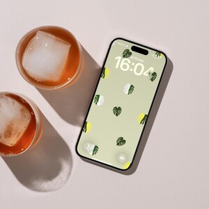 Op de afbeelding: Een smartphone met een lichtgroene achtergrond met een patroon van kleine monstera-bladeren in donkergroen, wit en geel. Het scherm toont de tijd 16:04 en de datum Za 22 aug. Twee glazen ijsthee staan in de buurt van het toestel.