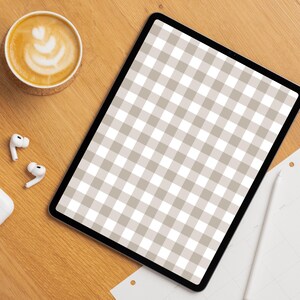 Op de afbeelding: Een digitale tablet met een beige en wit ruitpatroon op het scherm. Een kopje koffie met latte art op een onderzetter. Draadloze oortelefoons en een stylus zijn ook zichtbaar.