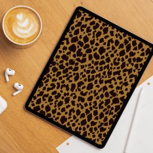 Puede incluir: Una tableta digital con un diseño de estampado de leopardo en tonos marrón y negro. La tableta está sobre una superficie de madera, junto a una taza de café y auriculares inalámbricos.