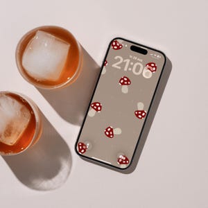 Op de afbeelding: Een smartphone met een digitale achtergrond met een patroon van rode en witte paddenstoelen op een beige achtergrond. Het scherm toont de tijd 21:06 en de datum Vr 28 aug. Twee glazen gevuld met amberkleurige vloeistof en grote ijsblokjes.