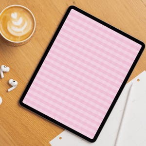 Op de afbeelding: Een tablet met een zwarte rand die een lichtroze en wit horizontaal gestreept patroon op het scherm weergeeft. Het apparaat ligt op een houten oppervlak naast een kop koffie, witte draadloze oordopjes en een witte styluspen.