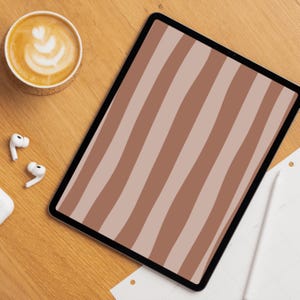 Op de afbeelding: Een digitale tablet met een bruin en beige gestreept patroon, een kop koffie met latte art, draadloze oordopjes en een stylus op een houten oppervlak.