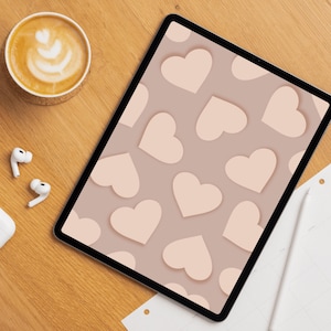 Op de afbeelding: Een digitale tablet toont een patroon van lichtroze harten op een taupe achtergrond. Een kopje koffie met latte art, witte oortelefoons en een stylus liggen op een houten oppervlak.