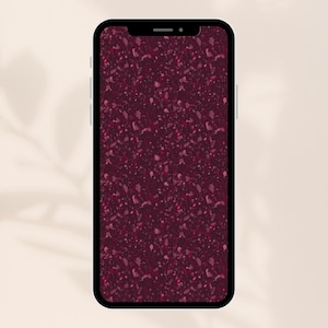 Op de afbeelding: Een smartphone met een zwarte rand toont een bordeauxrood terrazzo patroon. Het ontwerp van het scherm heeft een gespikkeld patroon van verschillende tinten rood en roze, waardoor een textuur ontstaat. De telefoon staat tegen een neutrale achtergrond.
