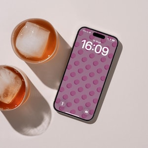 Op de afbeelding: Een smartphone met een vergrendelscherm met een paars stippenpatroon, waarop de tijd 16:09 en de datum Vr 10 jul wordt weergegeven, naast twee ronde glazen gevuld met amberkleurige vloeistof en grote vierkante ijsblokjes.