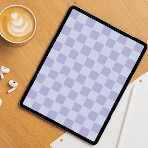 Op de afbeelding: Een tablet met een lichtpaars en lavendelkleurig ruitpatroon op het scherm. Het apparaat ligt op een houten ondergrond naast een kop koffie met latte art, witte draadloze oordopjes en een witte stylus op een papieren planner.