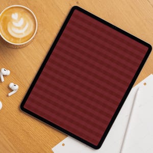 Op de afbeelding: Een tablet met een donkerrood en kastanjebruin horizontaal gestreept scherm, geplaatst naast een witte keramische beker met latte art, een paar witte draadloze oordopjes en een witte digitale stylus op een papieren planner.