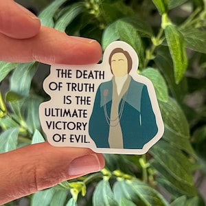 May include: A sticker featuring a teal and beige illustration of a person with the text "THE DEATH OF TRUTH IS THE ULTIMATE VICTORY OF EVIL." The sticker is held by a hand against a background of green leaves.
