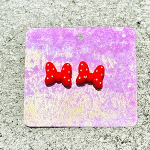 May include: A pair of red bow-shaped stud earrings with white polka dots. The earrings are displayed on a purple card with a small hole at the top. The background is a textured, light gray surface.