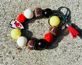 May include: A red, white and yellow beaded keychain with a football charm and a Kansas City Chiefs logo charm. The keychain also has a silver key ring and a red tassel.