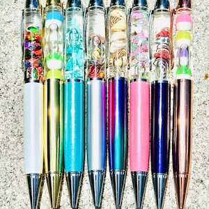 May include: Eight colorful pens with clear barrels filled with various objects, including seashells, glitter, and small toys. The pens have silver, gold, blue, silver, teal, pink, purple, and rose gold colored barrels.