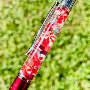 May include: A red and silver pen with a clear barrel filled with red and white candy-shaped decorations. The pen has a silver clip and tip, and the background is blurred green.