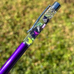 May include: A purple pen with a silver clip and a clear barrel filled with colorful, decorative elements. The pen's tip is black, and the barrel has a metallic sheen. The pen is held against a blurred green background.