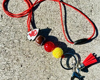 May include: Red, yellow and white beaded lanyard with a football charm and a red tassel keychain. The lanyard has a black breakaway clasp. The charm features the Kansas City Chiefs logo.
