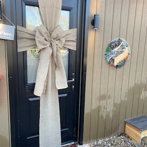 May include: A black door adorned with a large tan burlap bow and streamers. The door is set in a light brown wooden exterior. A sign that says "Welcome to my happy place" is to the left of the door.