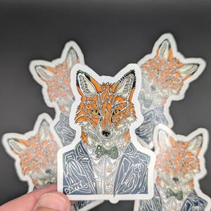 Puede incluir: Una pegatina troquelada que muestra un zorro naranja y blanco con una chaqueta de traje azul oscuro, una camisa blanca y una pajarita verde. El diseño incluye líneas intrincadas, la palabra DAPPER y el texto FOX 2024 integrado en la ilustración.
