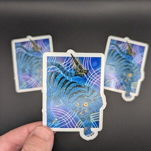 以下が含まれることがあります： 黒い縞模様と輝く黄色い目を持つ青い虎が描かれた長方形のステッカー。虎の頭には角があり、青と紫のグラデーションの背景に、白とピンクの曲線が交差するデザインが施されています。