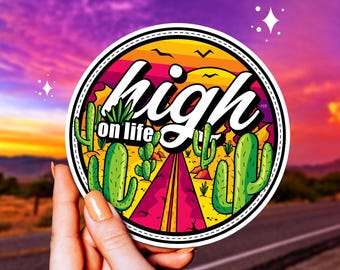 May include: A round sticker with a desert scene and the text "high on life" in white lettering. The sticker has a pink, yellow, and orange color scheme with a road leading through the desert.