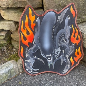 May include: A black leather patch featuring a detailed illustration of an alien creature with flames on either side. The alien is grey and has a large head with its mouth open in a menacing expression.