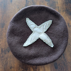 May include: A brown wool beret with a large, off-white X stitched on the front. The X is made of a lighter fabric and outlined with a contrasting stitch. The beret is round and has a soft texture.