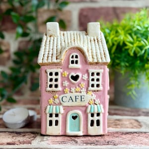 May include: A ceramic house-shaped tea light holder in pink with a white textured roof. The facade features a central door, multiple windows, a sign reading CAFE, and small yellow and white floral accents. A heart-shaped cutout sits above the sign.